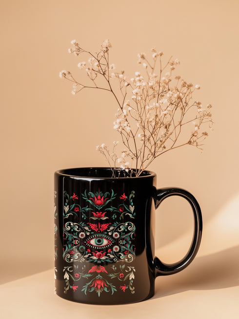 A black ceramic mug featuring a symmetrical folk art design with a central mystic eye motif in turquoise and coral. The design includes ornate scrollwork, red tulip-like flowers, sage green leaves, and cream flourishes arranged in a decorative pattern. The mug has a glossy finish with a C-shaped handle.